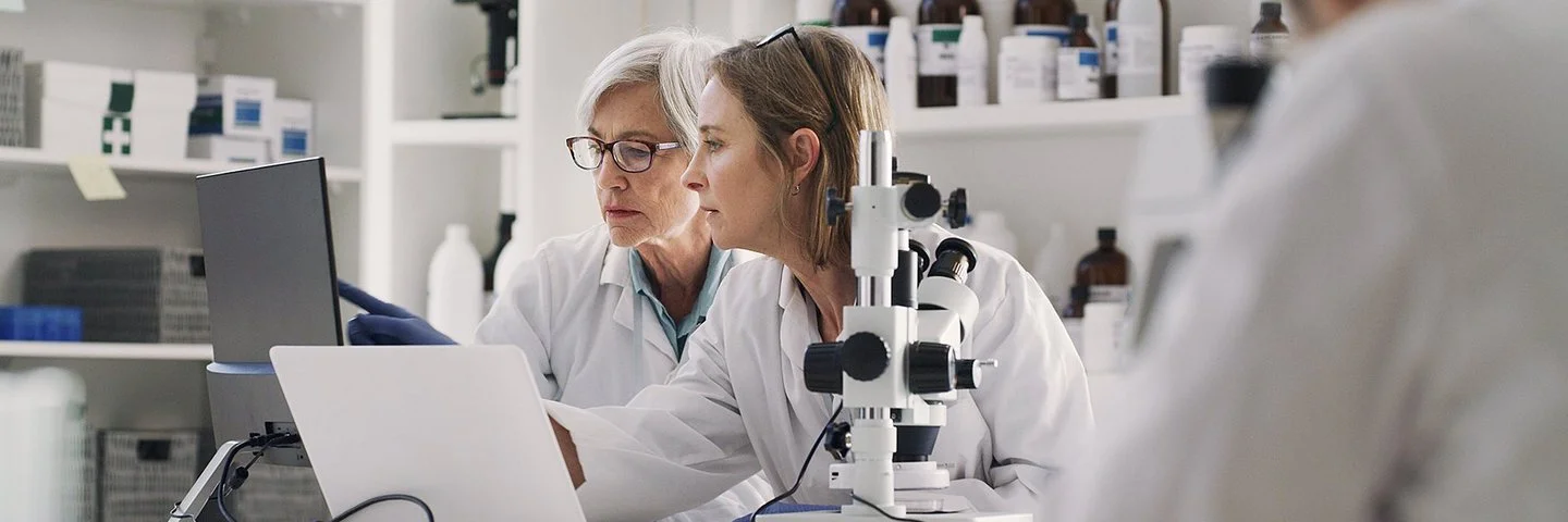 In einem medizinischen Labor sitzen zwei Frauen in weißen Kitteln an einem Arbeitstisch. Auf dem Tisch stehen ein Mikroskop, ein aufgeklappter Laptop und ein Monitor. Beide schauen auf den Monitor, auf dem eine der Frauen mit dem Zeigefinger auf etwas zeigt.
