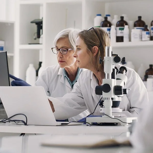 In einem medizinischen Labor sitzen zwei Frauen in weißen Kitteln an einem Arbeitstisch. Auf dem Tisch stehen ein Mikroskop, ein aufgeklappter Laptop und ein Monitor. Beide schauen auf den Monitor, auf dem eine der Frauen mit dem Zeigefinger auf etwas zeigt.