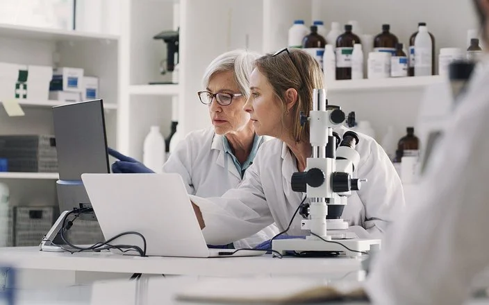 In einem medizinischen Labor sitzen zwei Frauen in weißen Kitteln an einem Arbeitstisch. Auf dem Tisch stehen ein Mikroskop, ein aufgeklappter Laptop und ein Monitor. Beide schauen auf den Monitor, auf dem eine der Frauen mit dem Zeigefinger auf etwas zeigt.