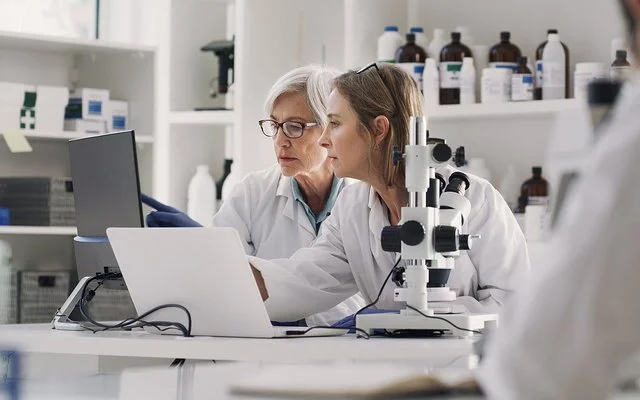 In einem medizinischen Labor sitzen zwei Frauen in weißen Kitteln an einem Arbeitstisch. Auf dem Tisch stehen ein Mikroskop, ein aufgeklappter Laptop und ein Monitor. Beide schauen auf den Monitor, auf dem eine der Frauen mit dem Zeigefinger auf etwas zeigt.