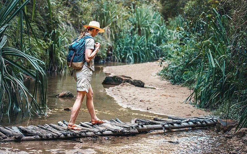 Eine Frau in kurzer Freizeitkleidung läuft in den Tropen über einen Holzsteg auf sandiges Ufer zu. An den Füßen trägt sie Sandalen.