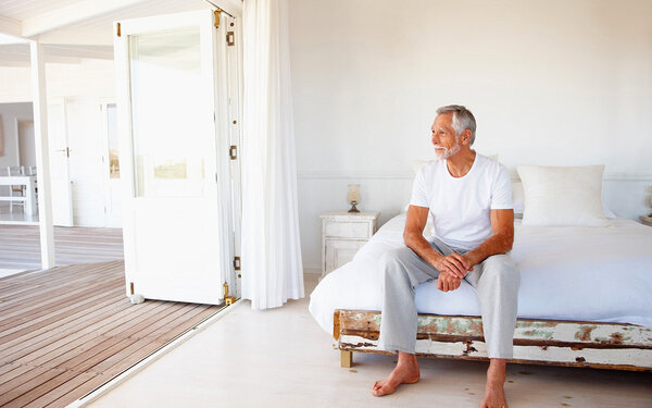 Ein Mann mit Muskelschwund im Alter sitzt auf der Bettkante und blickt durch die geöffnete Terassentür.