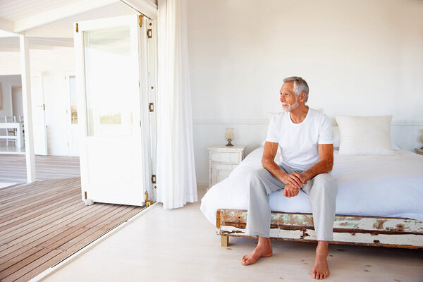 Ein Mann mit Muskelschwund im Alter sitzt auf der Bettkante und blickt durch die geöffnete Terassentür.