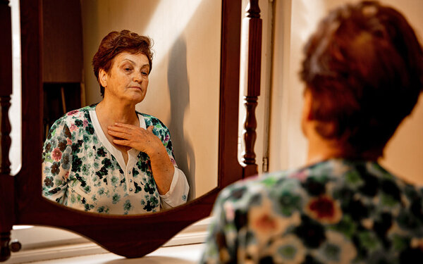 Eine Seniorin schaut skeptisch in den Spiegel, während sie mit der rechten Hand die Haut an ihrem Hals betastet.