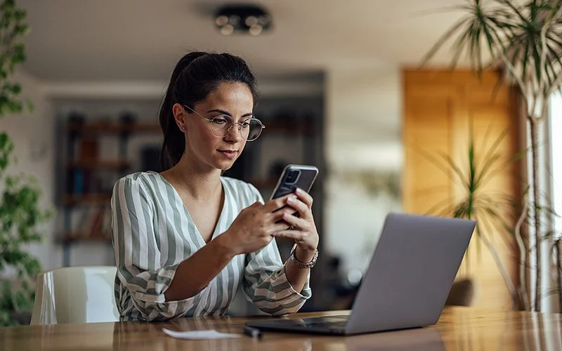 Eine junge Frau sitzt an einem Tisch im Wohnzimmer. Vor ihr auf dem Tisch steht ein aufgeklappter Laptop. In den Händen hält sie ein Smartphone und liest etwas auf dessen Display.