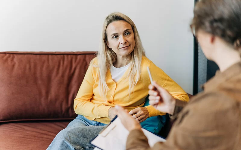 Im Sprechzimmer einer Psychotherapeutin sitzt eine Frau auf dem Sofa und hört interessiert den Ausführungen der Therapeutin zu.