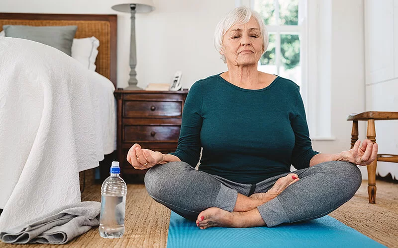 Eine Seniorin meditiert in der Lotusposition auf dem Boden in ihrem Schlafzimmer.