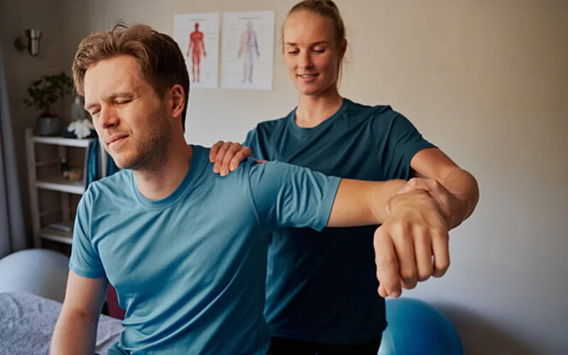 Ein junger Mann ist bei einer Physiotherapeutin, die ihm mit Krankengymnastik bei den Beschwerden in der Schulter hilft.