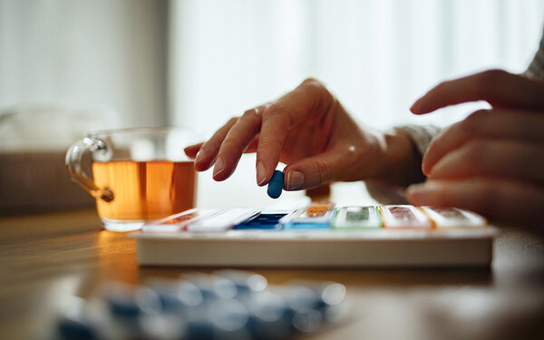 Hände entnehmen eine blaue Pille aus einer bunten Medikamentendose.