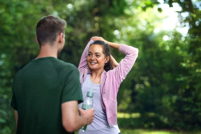 Eine junge Frau in den Zwanzigern steht in einem Park und mach Dehnübungen mit den Armen. Ihr gegenüber steht ein nicht erkennbarer Mann mit einer Trinkflasche in der rechten Hand.