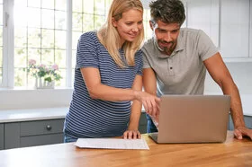 Ein Paar mit schwangerer Frau schaut gemeinsam auf den Bildschirm eines Laptops.