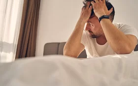 Ein Mann mit Geschlechtskrankheiten sitzt auf dem Bett, fasst sich an den Kopf und blickt nachdenklich aus dem Fenster.