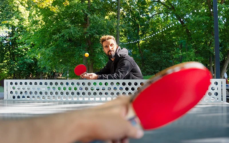 Ein Tischtennisspiel auf einem Freiluftsportplatz. Eine der spielenden Personen ist nicht vollständig im Bild, sondern nur der Unterarm mit einem Schläger in der Hand. Am anderen Ende der Tischtennisplatte steht ein Mann, der zum Zurückspielen bereit ist, während der Tischtennisball auf ihn zufliegt.