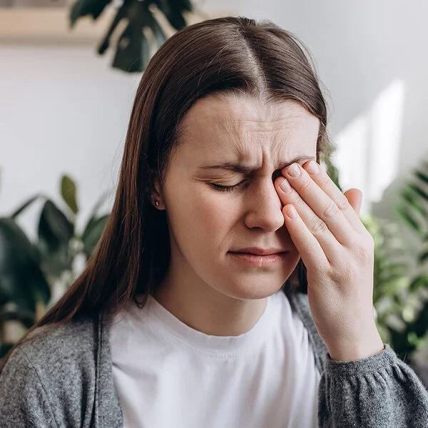 Eine junge Frau sitzt mit schmerzerfülltem Gesicht in einem Wohnzimmer. Ihre Augen sind geschlossen. Mit der linken Hand fasst sie sich an das linke Auge.