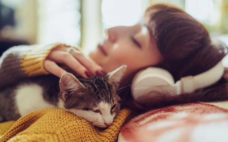 Eine junge Frau mit Skin-Picking-Disorder, die entspannt auf der Couch liegt, mit Kopfhörern Musik hört und dabei mit einer gefleckten Katze kuschelt.