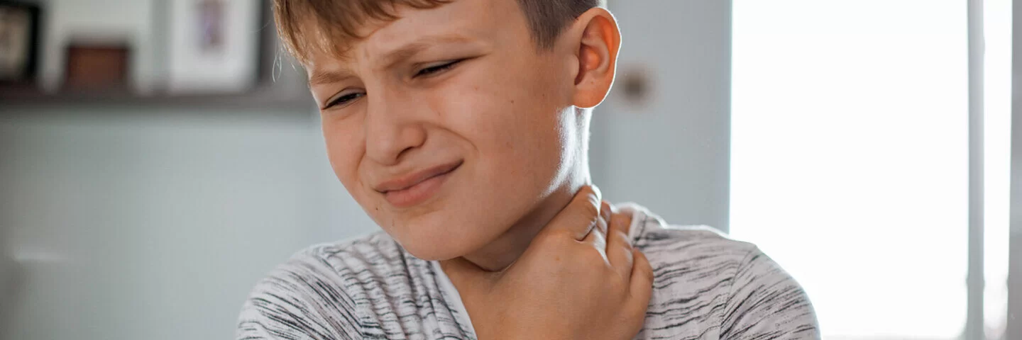 Ein Junge, der eine Mandelentzündung hat, fasst sich mit schmerzverzerrtem Gesicht an den Hals.