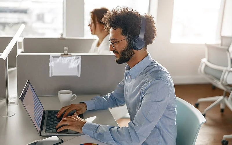 In einem Großraumbüro sitzen ein Mann und eine Frau an ihren Schreibtischen. Der Mann, der im Vordergrund sitzt, tippt in einen Laptop und trägt Kopfhörer.