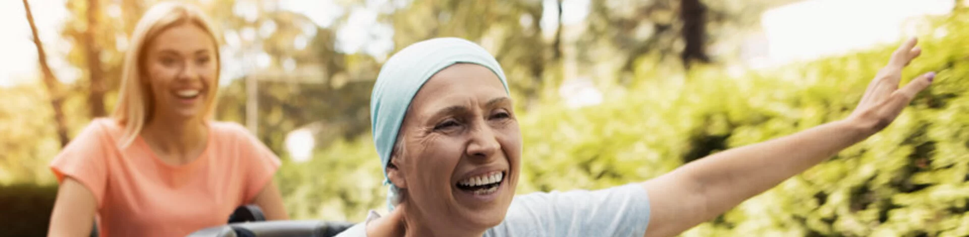 Eine lachende Frau schiebt eine Lungenkrebs-Patientin im Rollstuhl, die glücklich die Arme nach oben streckt.