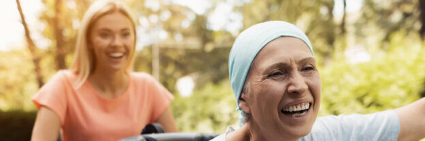 Eine lachende Frau schiebt eine Lungenkrebs-Patientin im Rollstuhl, die glücklich die Arme nach oben streckt.