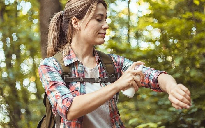 Eine Frau mit Rucksack sitzt auf einem Baumstamm im Wald und sprüht sich die nackte Haut der Unterarme mit Insektenschutzmittel ein.