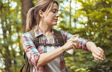 Eine Frau mit Rucksack sitzt auf einem Baumstamm im Wald und sprüht sich die nackte Haut der Unterarme mit Insektenschutzmittel ein.