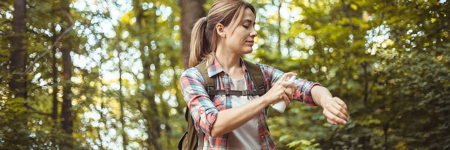 Eine Frau mit Rucksack sitzt auf einem Baumstamm im Wald und sprüht sich die nackte Haut der Unterarme mit Insektenschutzmittel ein.