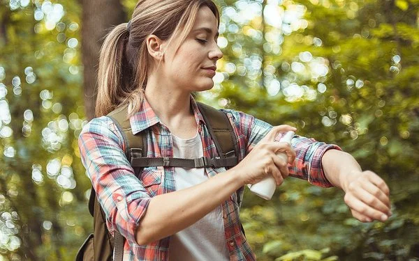 Eine Frau mit Rucksack sitzt auf einem Baumstamm im Wald und sprüht sich die nackte Haut der Unterarme mit Insektenschutzmittel ein.