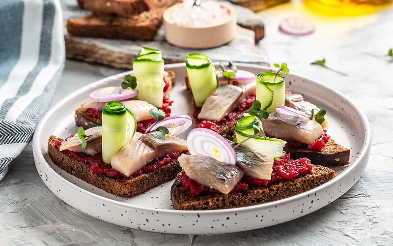 Auf einem Teller liegen mehrere Scheiben dunkles Brot. Sie sind mit Hering, Roter Bete, Gurke, Zwiebeln und Microgreens belegt.