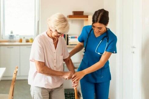 Pflegebedürftige Person stützt sich beim Gehen auf einen Stock und wird dabei von einer Pflegekraft unterstützt.