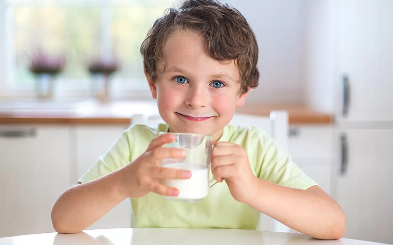 Ein kleiner Junge sitzt lächelnd am Küchentisch und hält ein Glas Milch in beiden Händen.