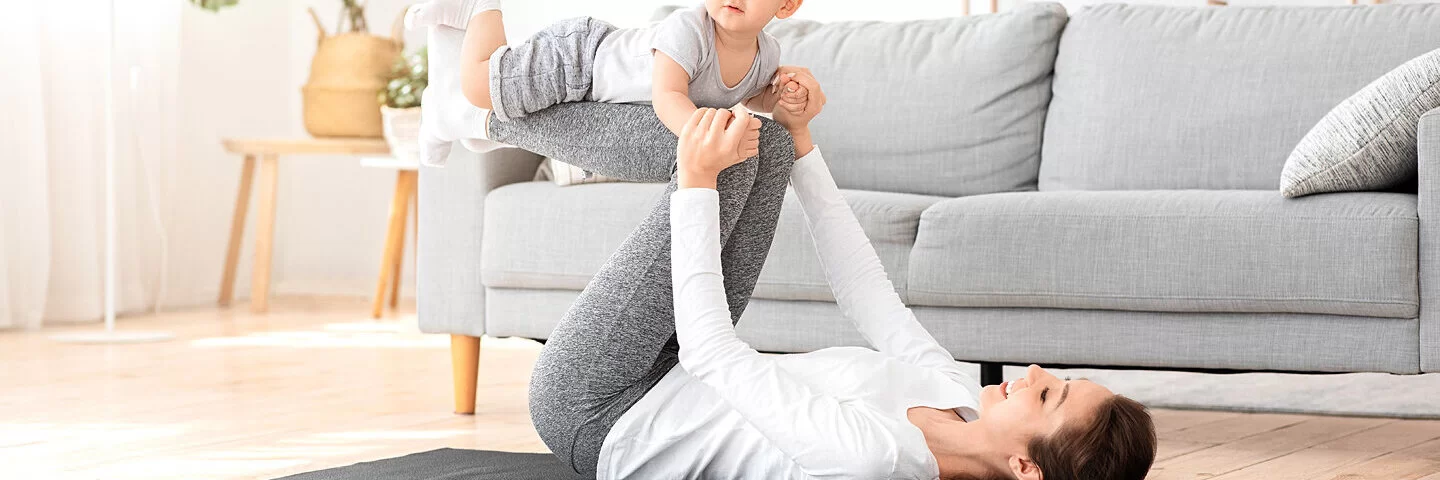 Eine junge Mutter macht Fitnessübungen und ihr Kind liegt auf ihren Beinen.