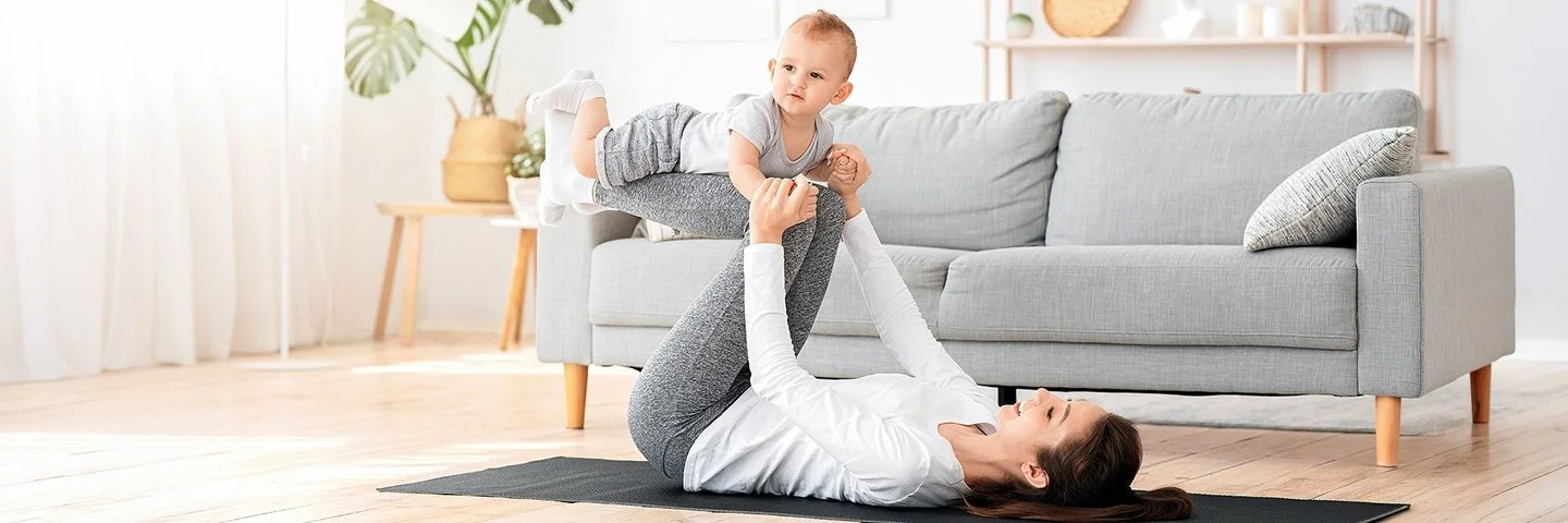 Eine junge Mutter macht Fitnessübungen und ihr Kind liegt auf ihren Beinen.