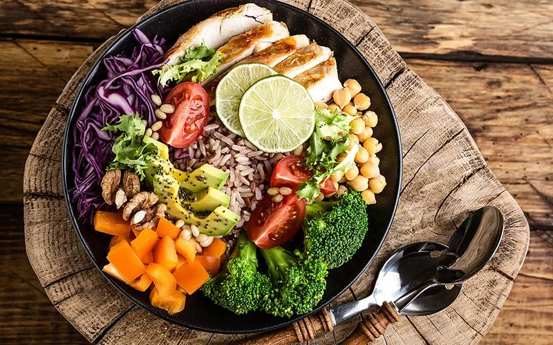 Obenansicht einer Salatschüssel mit Hähnchenfilet, Naturreis, Avocado, Pfeffer, Tomaten, Brokkoli, Rotkohl, Kichererbsen, frischem Blattsalat, Pinienkernen und Walnüssen. Die schwarze Keramikschüssel steht auf einem Holzbrett, das sich wiederum auf einem Holztisch befindet.