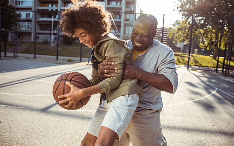 Ein Vater spielt mit seinem Sohn Basketball, beide haben Spaß an Bewegung.
