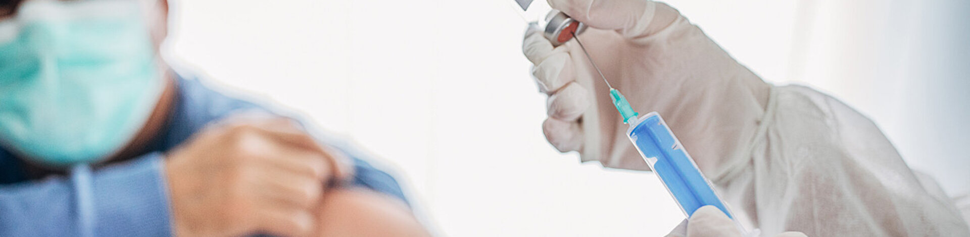 Ein Mann mit Mundschutz sitzt in einer Arztpraxis, im Vordergrund wird eine Spritze aufgezogen.