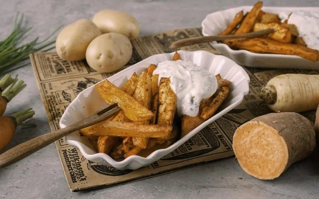 Weiße Schale mit Gemüsefritten, weißem Dip und Pommesgabel auf einem rustikalen Untergrund. Im Hintergrund Kartoffeln, Süßkartoffeln, Pastinaken und Möhren.