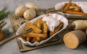 Weiße Schale mit Gemüsefritten, weißem Dip und Pommesgabel auf einem rustikalen Untergrund. Im Hintergrund Kartoffeln, Süßkartoffeln, Pastinaken und Möhren.