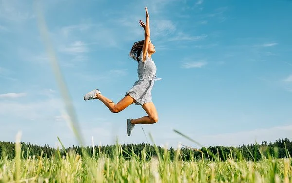 Eine Frau in kurzer Sommerkleidung springt fröhlich und ausgelassen über eine grüne Wiese bei strahlend blauem Himmel.