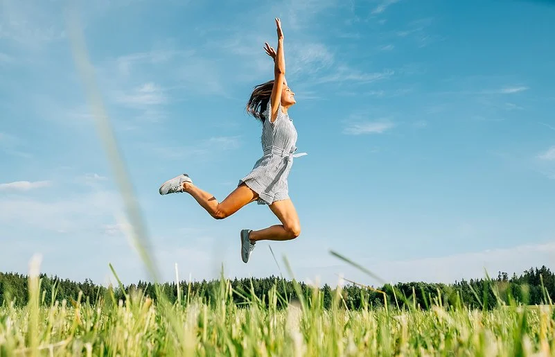 Eine Frau in kurzer Sommerkleidung springt fröhlich und ausgelassen über eine grüne Wiese bei strahlend blauem Himmel.