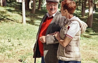Zwei Personen gehen im Park, der Senior stützt sich auf einen Gehstock und die Frau hält den Arm des Mannes.
