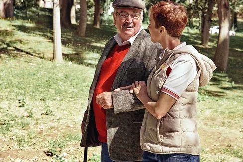 Zwei Personen gehen im Park, der Senior stützt sich auf einen Gehstock und die Frau hält den Arm des Mannes.