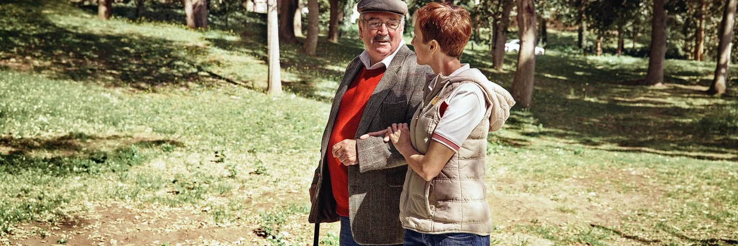 Zwei Personen gehen im Park, der Senior stützt sich auf einen Gehstock und die Frau hält den Arm des Mannes.