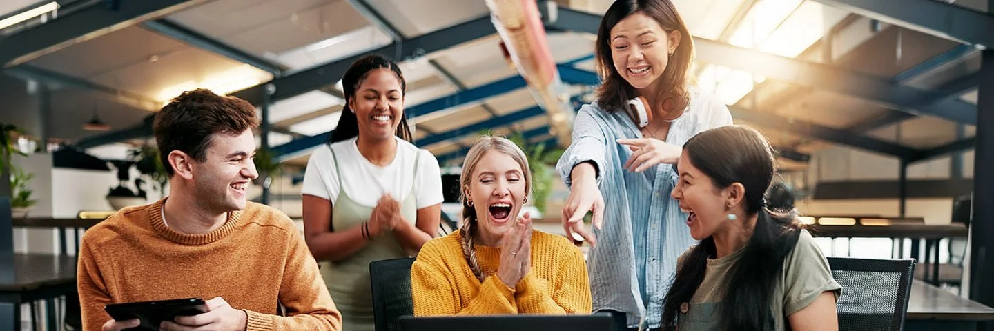Eine Gruppe junger Menschen sitzt lachend um einen Laptop an einem Tisch in einem Büro.