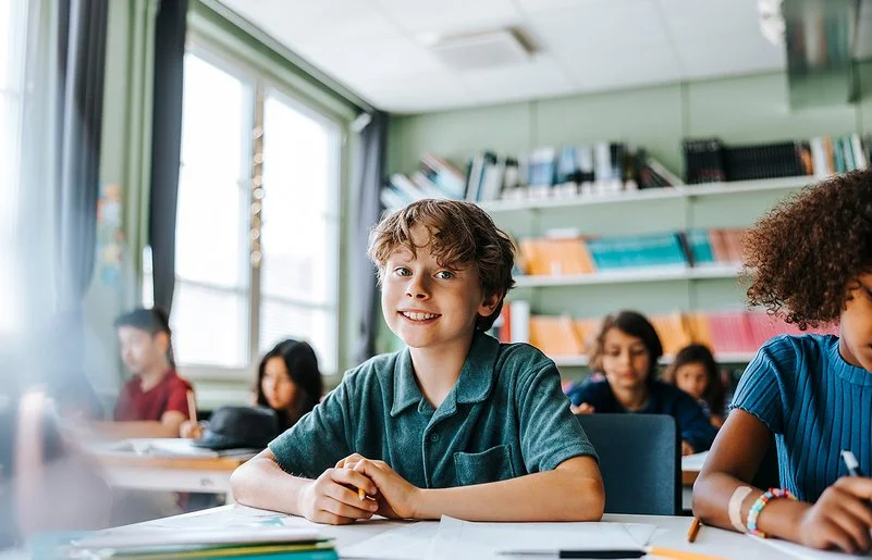 Ein Junge mit lockigem Haar und einem blauen Kurzarmhemd sitzt in einem Klassenzimmer und lächelt in die Kamera.