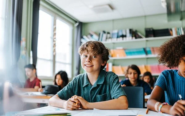 Ein Junge mit lockigem Haar und einem blauen Kurzarmhemd sitzt in einem Klassenzimmer und lächelt in die Kamera.