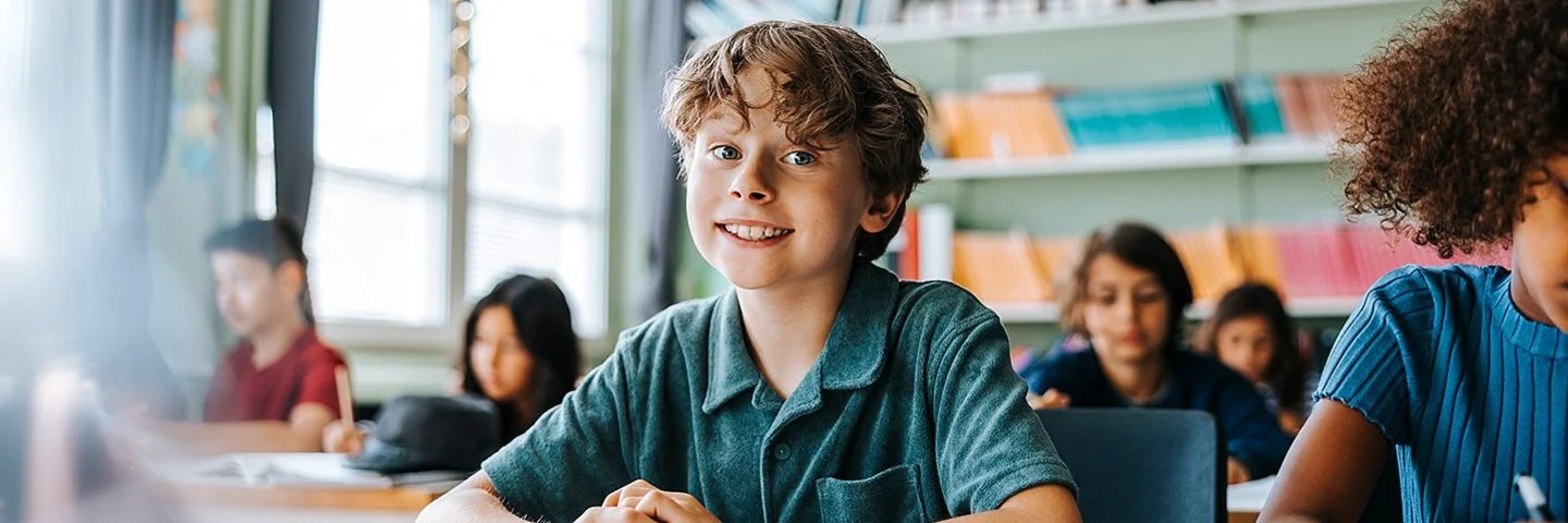 Ein Junge mit lockigem Haar und einem blauen Kurzarmhemd sitzt in einem Klassenzimmer und lächelt in die Kamera.