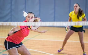 Eine Frau steht nah am Netz und schlägt einen Federball mit einem Badmintonschläger, während ihre Spielpartnerin, die hinter ihr steht, sie anschaut.