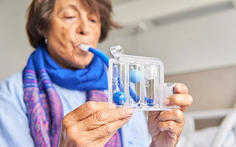 Nahaufnahme einer älteren Frau mit lila gemustertem Schal in einem Krankenzimmer. Sie benutzt einen Atemtrainer zur Förderung der Genesung nach einem Rippenbruch.