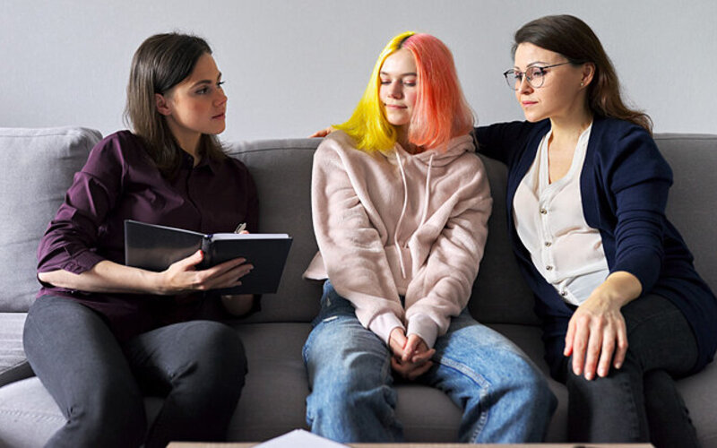 Eine junge Therapeutin bespricht sich auf einem Sofa mit einem psychisch kranken Teenager-Mädchen und ihrer Mutter.