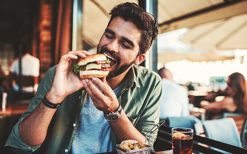 Junger Mann im Restaurant, der in einen Burger mit Fleisch beißt.